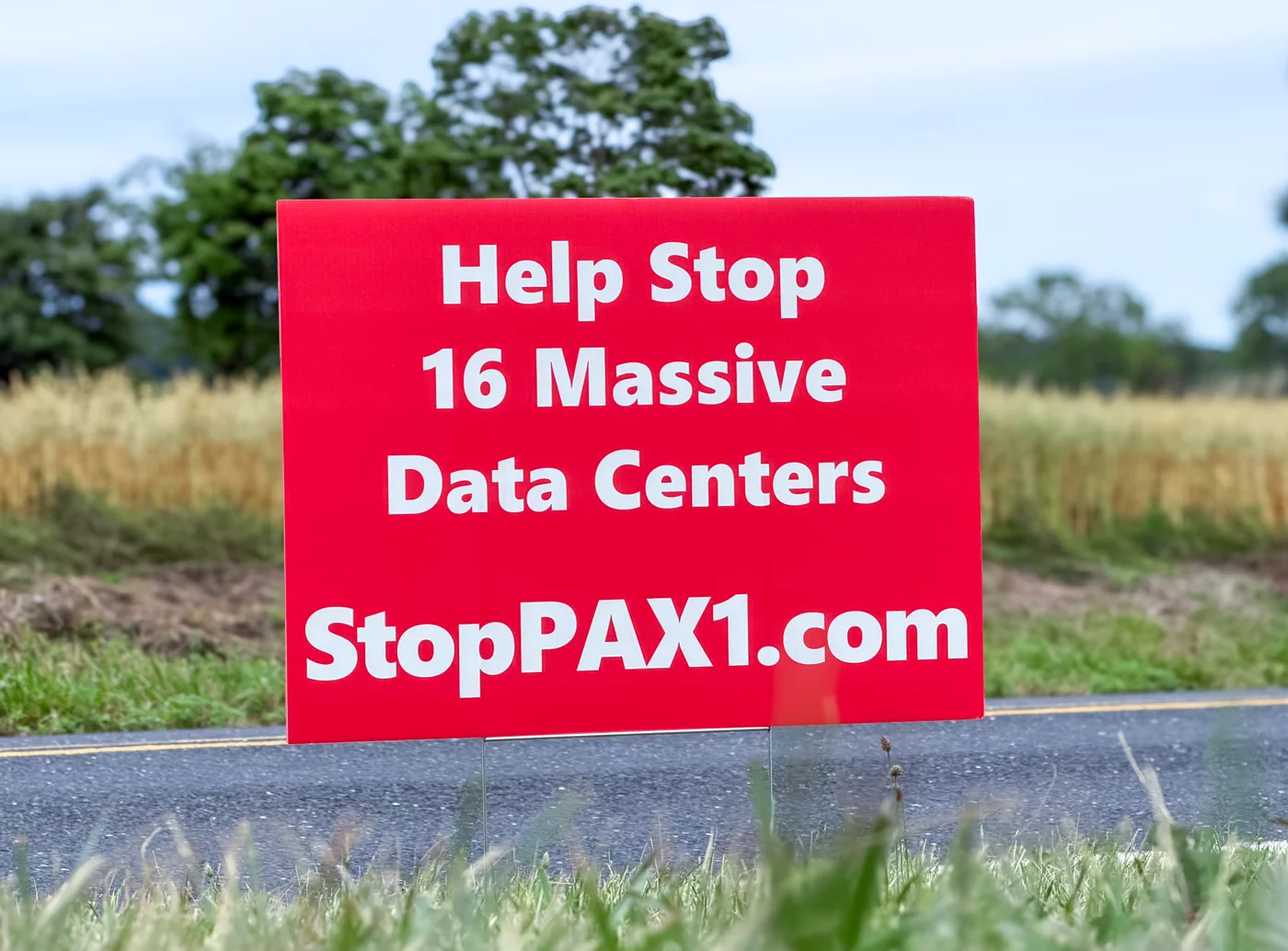 A red Stop PAX-1 yard sign at the edge of a road reading 'Help Stop 16 Massive Data Centers, StopPAX1.com', with farmland behind it.